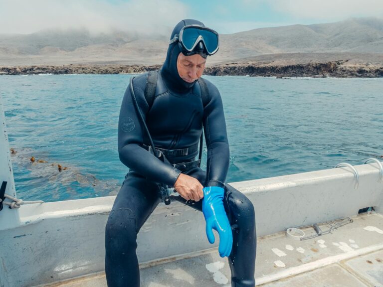 A scuba diver wearing a wetsuit prepares blue gloves on a boat with ocean view.